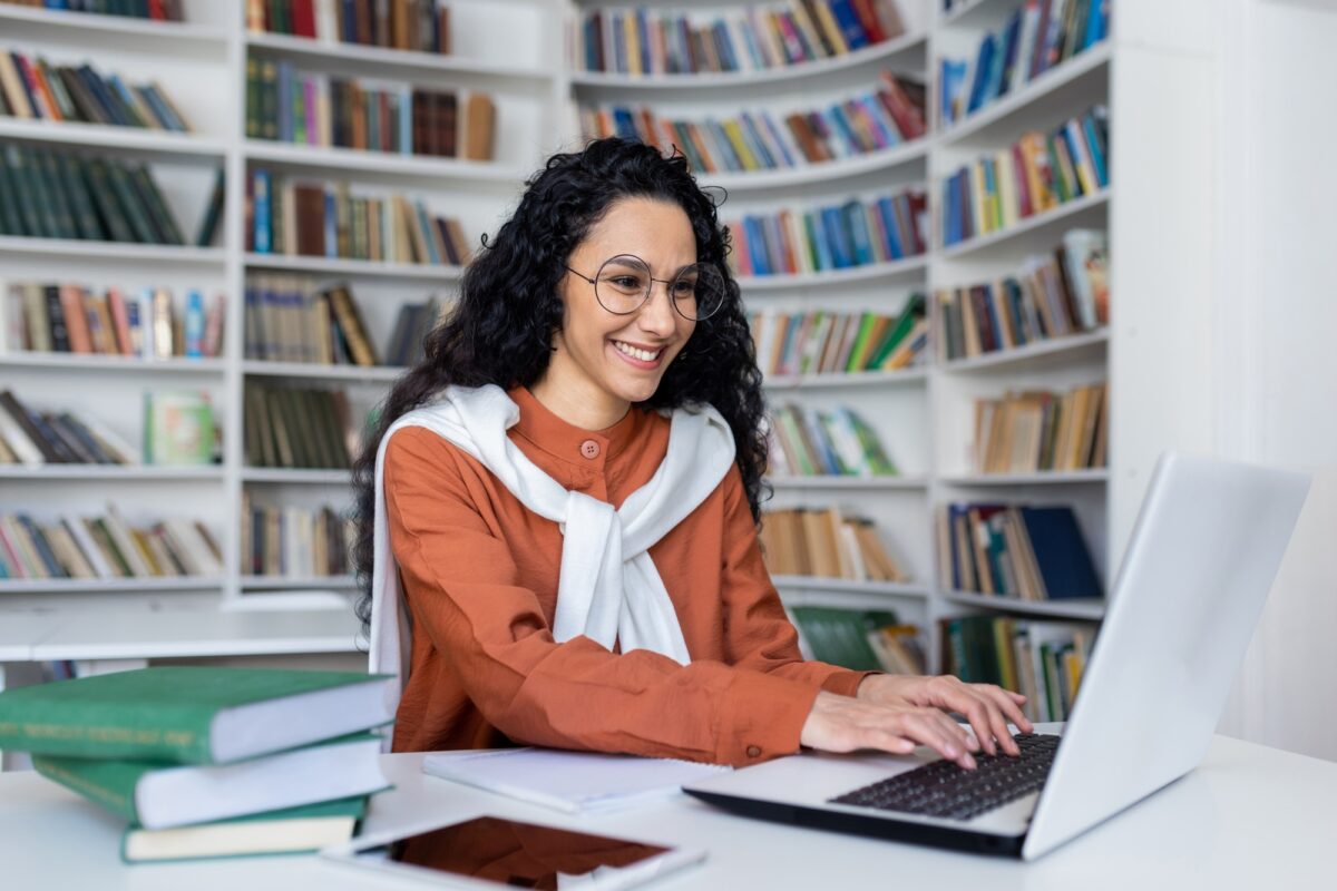 Smiling casual brunette typing on laptop keyboard while sitting in campus library with academical