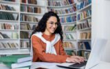Smiling casual brunette typing on laptop keyboard while sitting in campus library with academical
