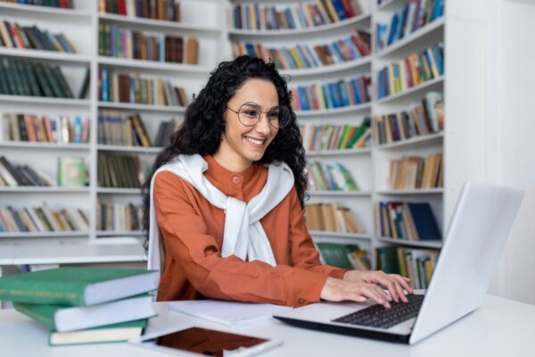 Smiling casual brunette typing on laptop keyboard while sitting in campus library with academical
