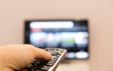 Watching TV and using remote controller. Hand holding TV remote control with a television in the
